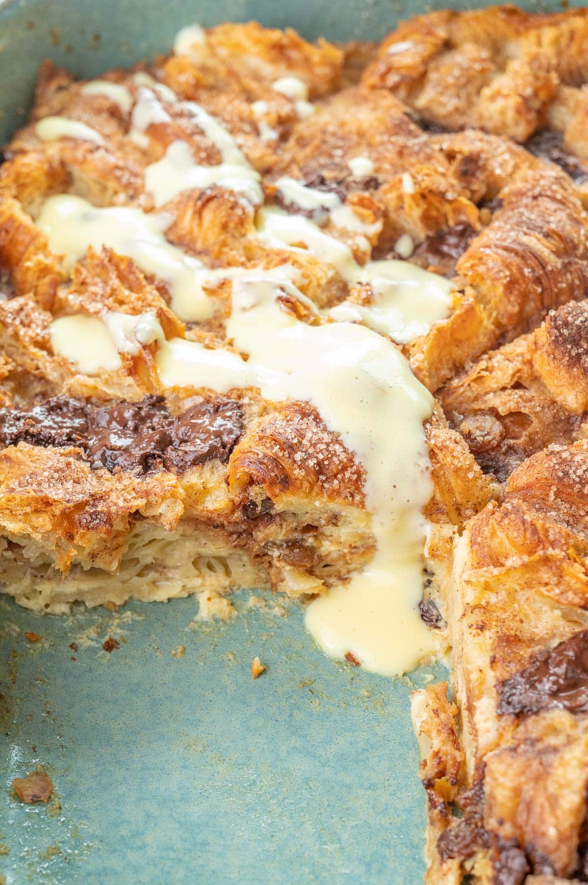 Croissant bread pudding in a baking dish with a piece missing, drizzled with vanilla sauce.
