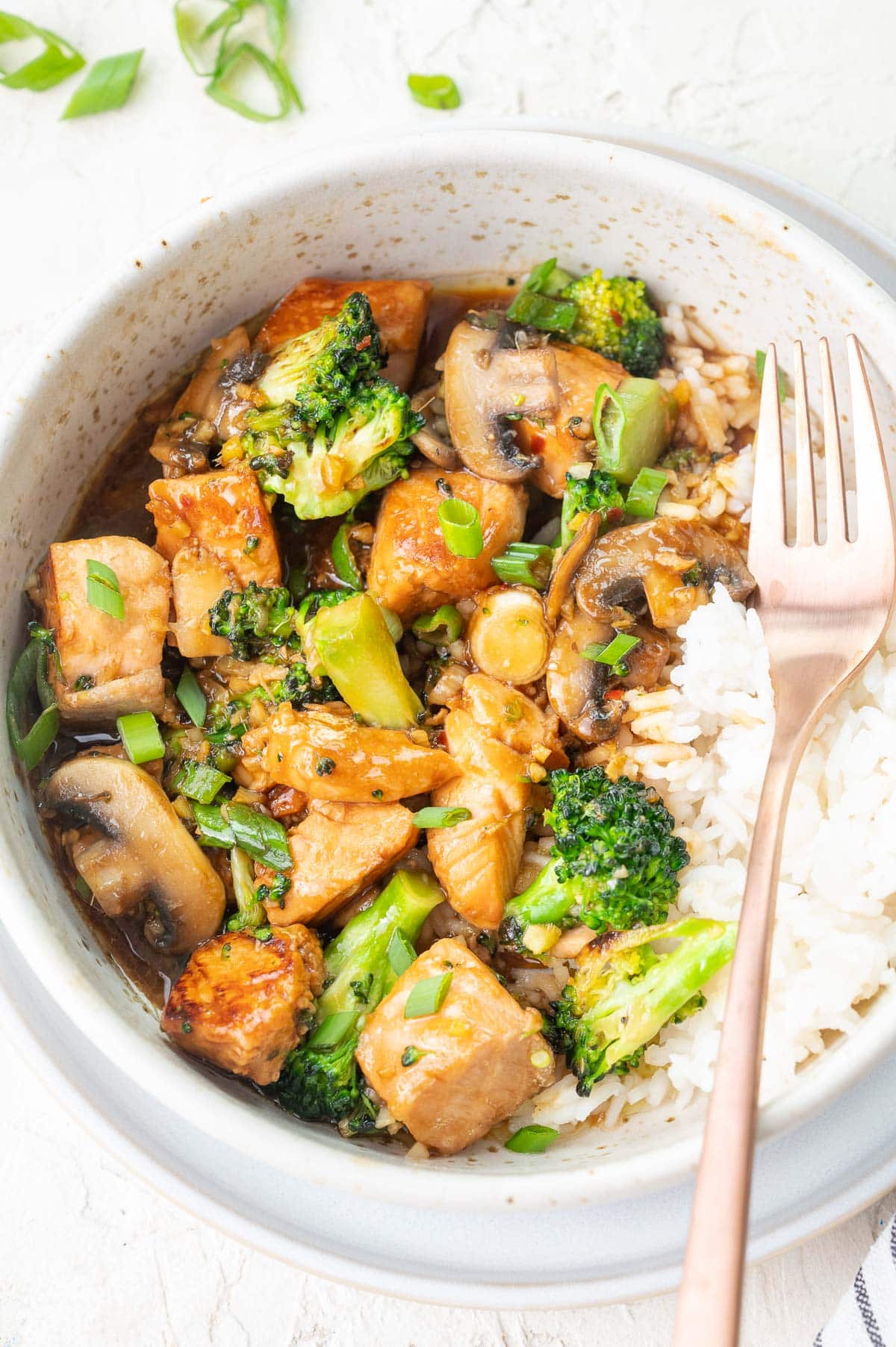 A close up photo of salmon stir fry with broccoli, mushrooms, and rice in a white bowl.