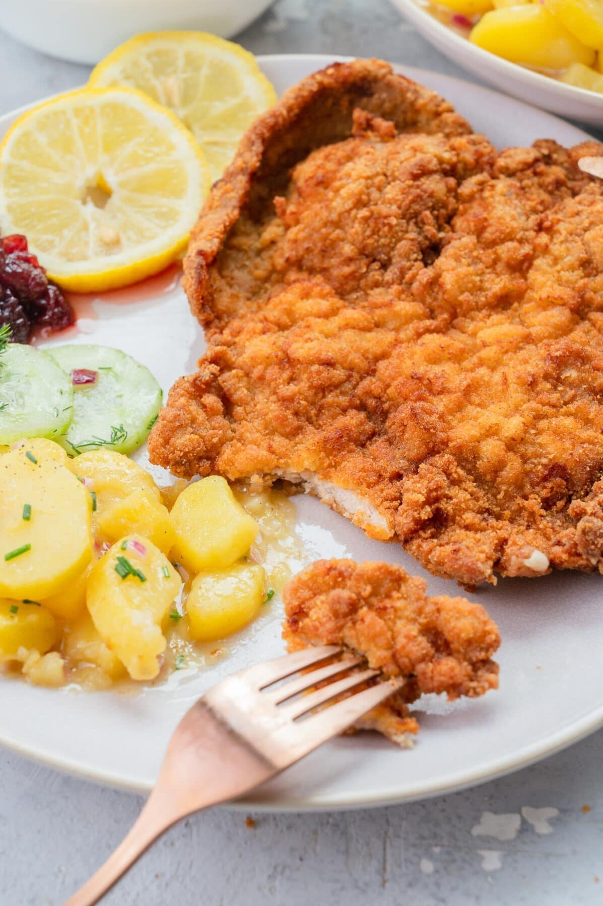 Wiener Schnitzel on a beige plate with lemon slices, potato and cucumber salad.