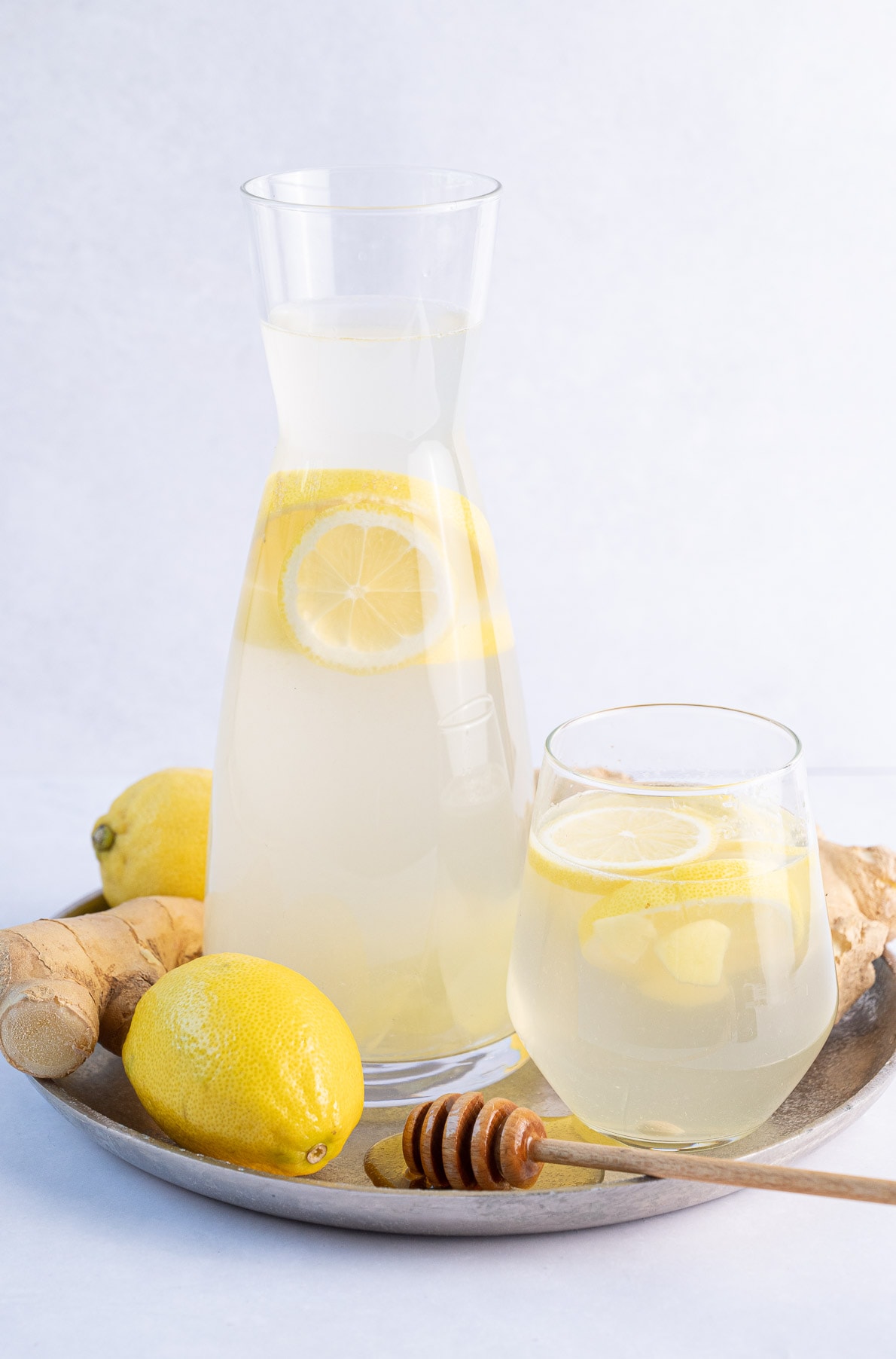 A pitcher and a glass with lemon ginger water, lemons, fresh ginger, and honey on a silver tray.