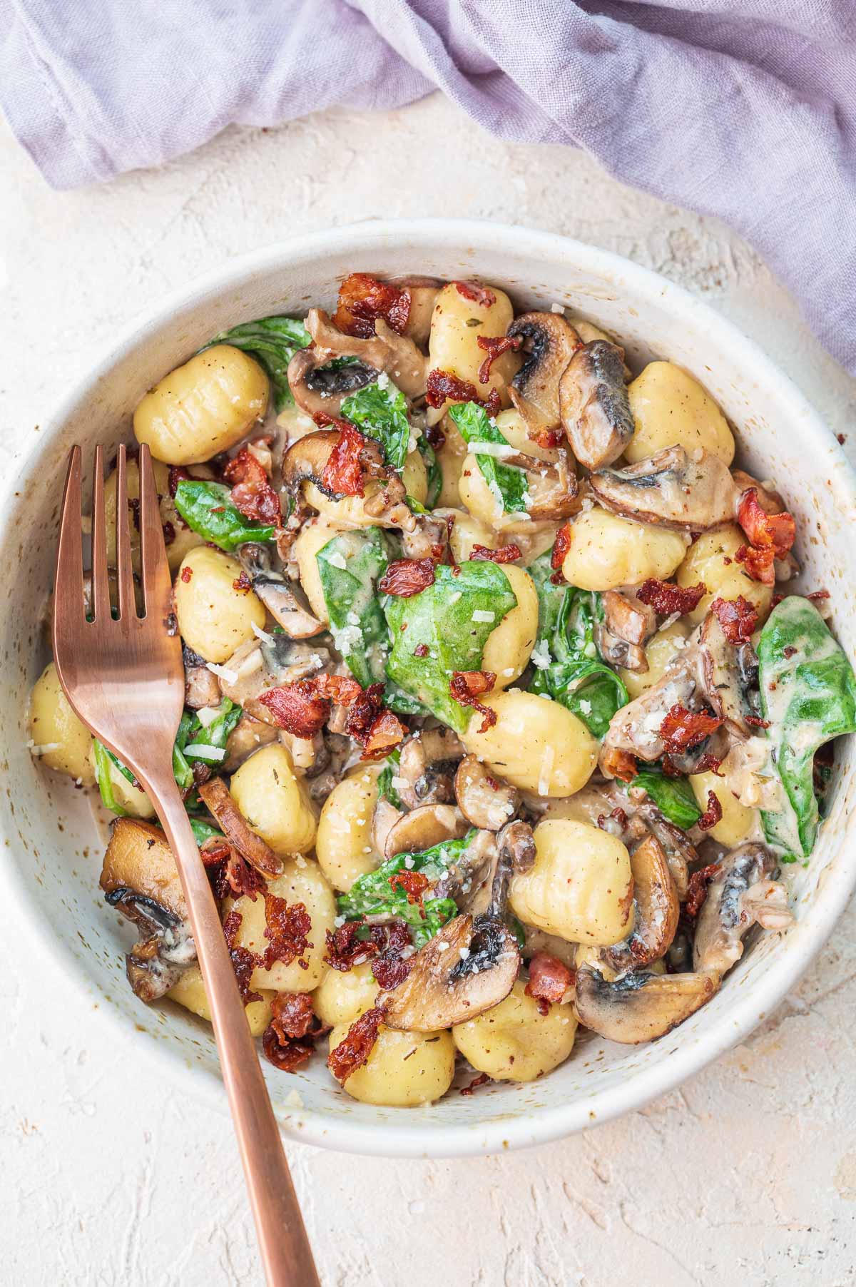 Mushroom gnocchi with spinach and bacon in a white bowl with a gold fork on a side.