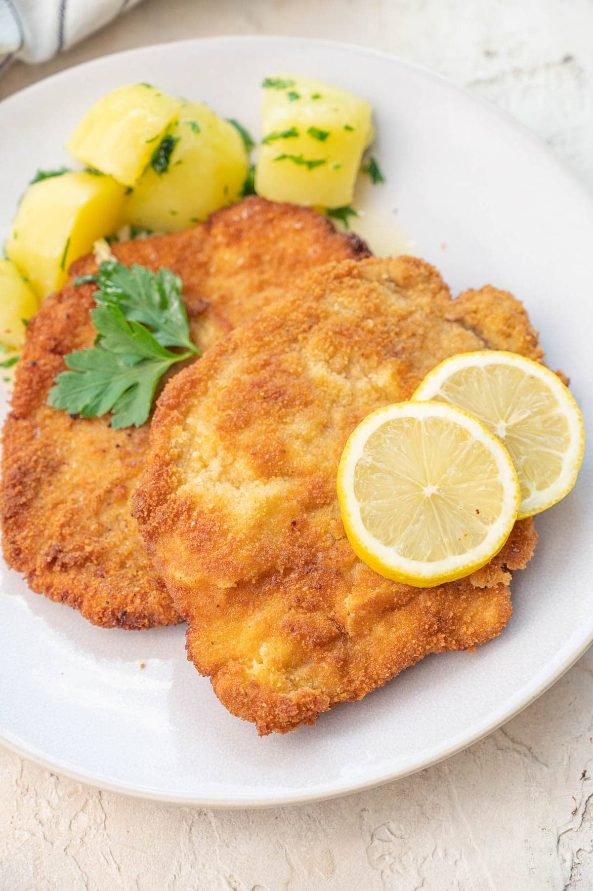German pork schnitzel topped with lemon slices and served with potatoes on a beige plate.