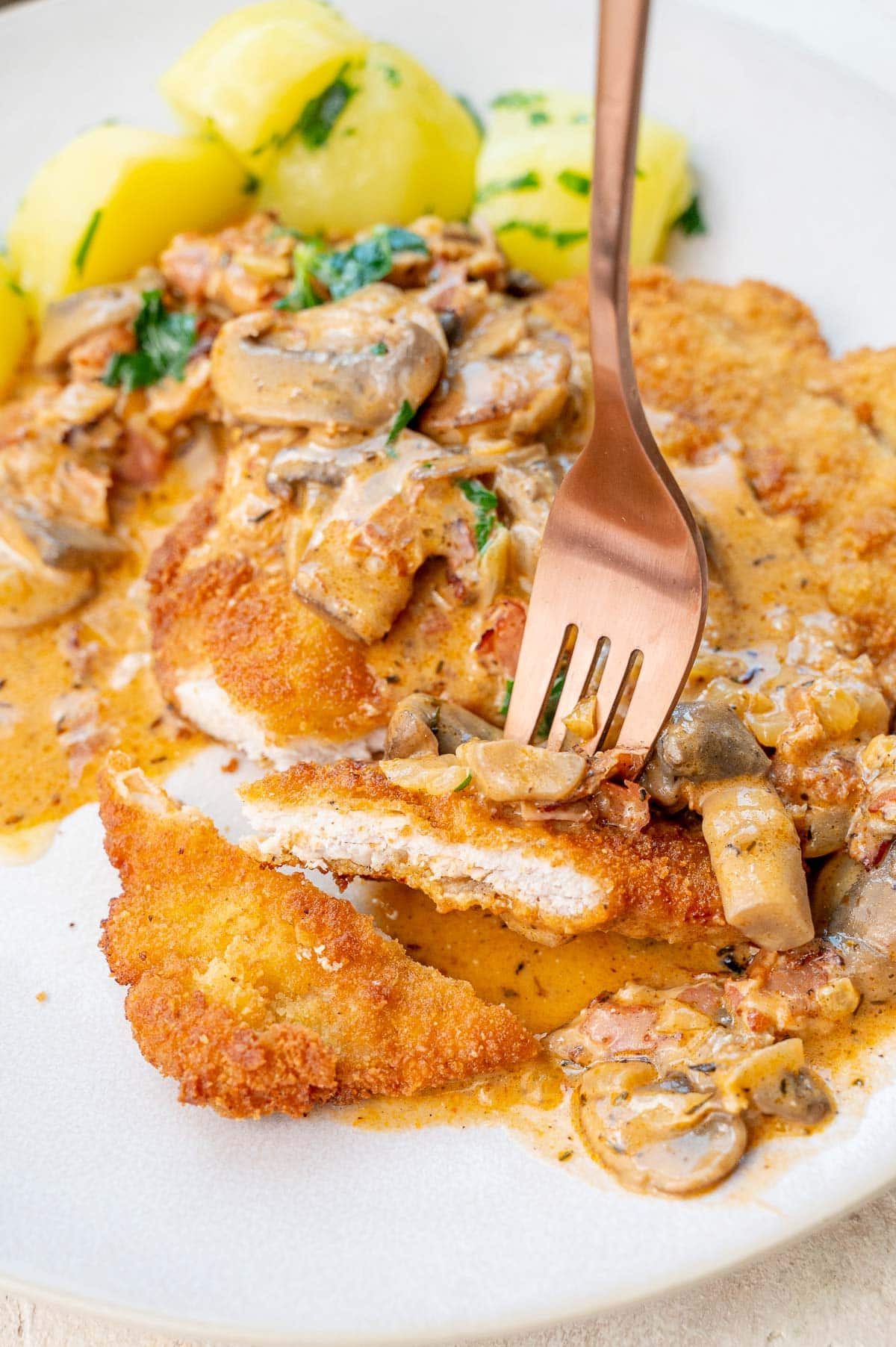 Jägerschnitzel with mushroom gravy and potatoes on a beige plate. A piece of the Schnitzel is stuck on a gold fork.
