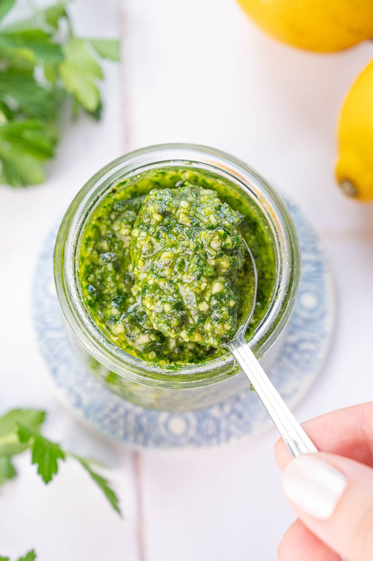 An overhead photo of parsley pesto in a jar being scooped with a teaspoon. Parsley leaves and lemons in the background.