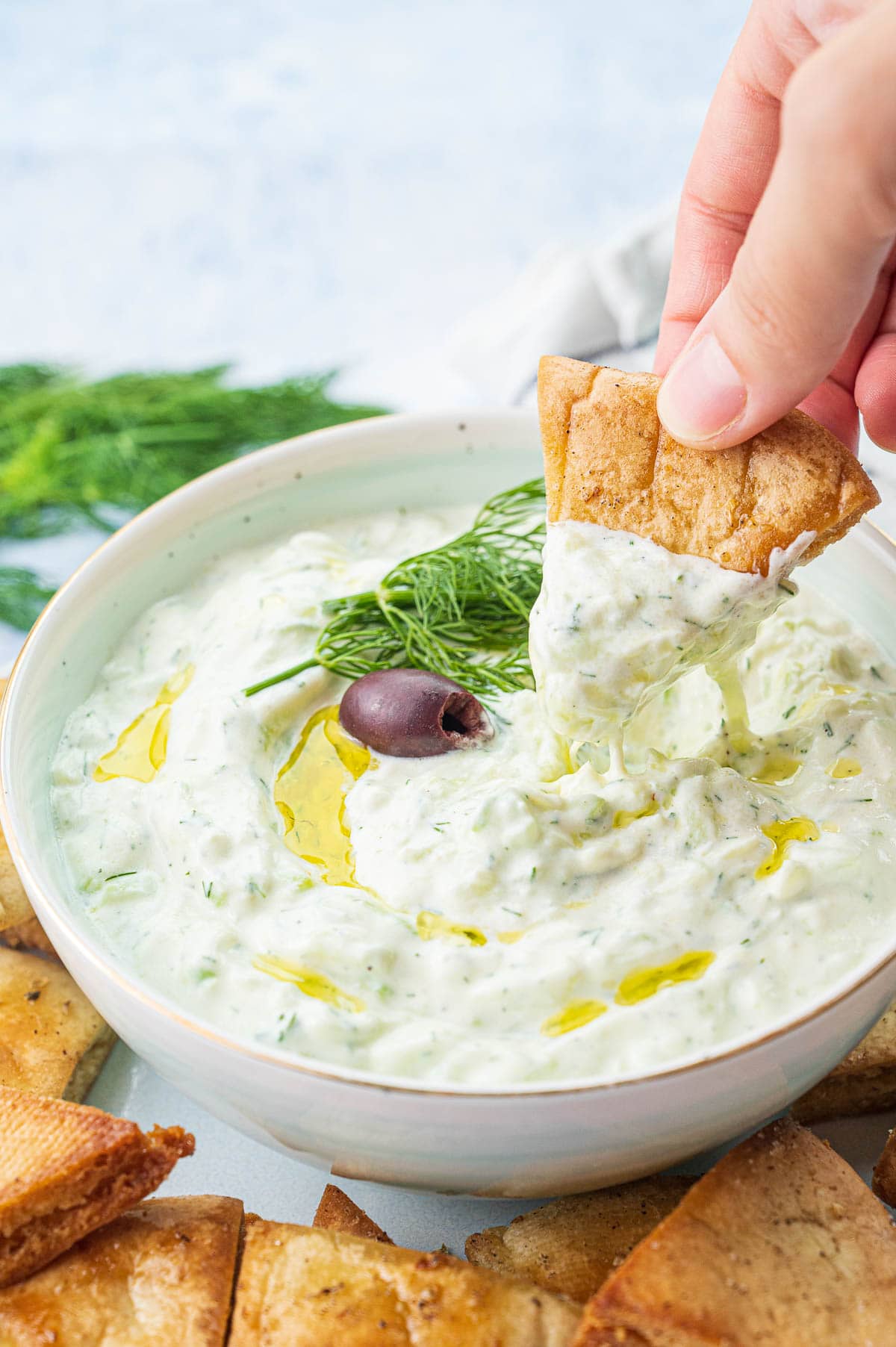 Tzatziki dip in a white bowl is being scooped with pita chips.