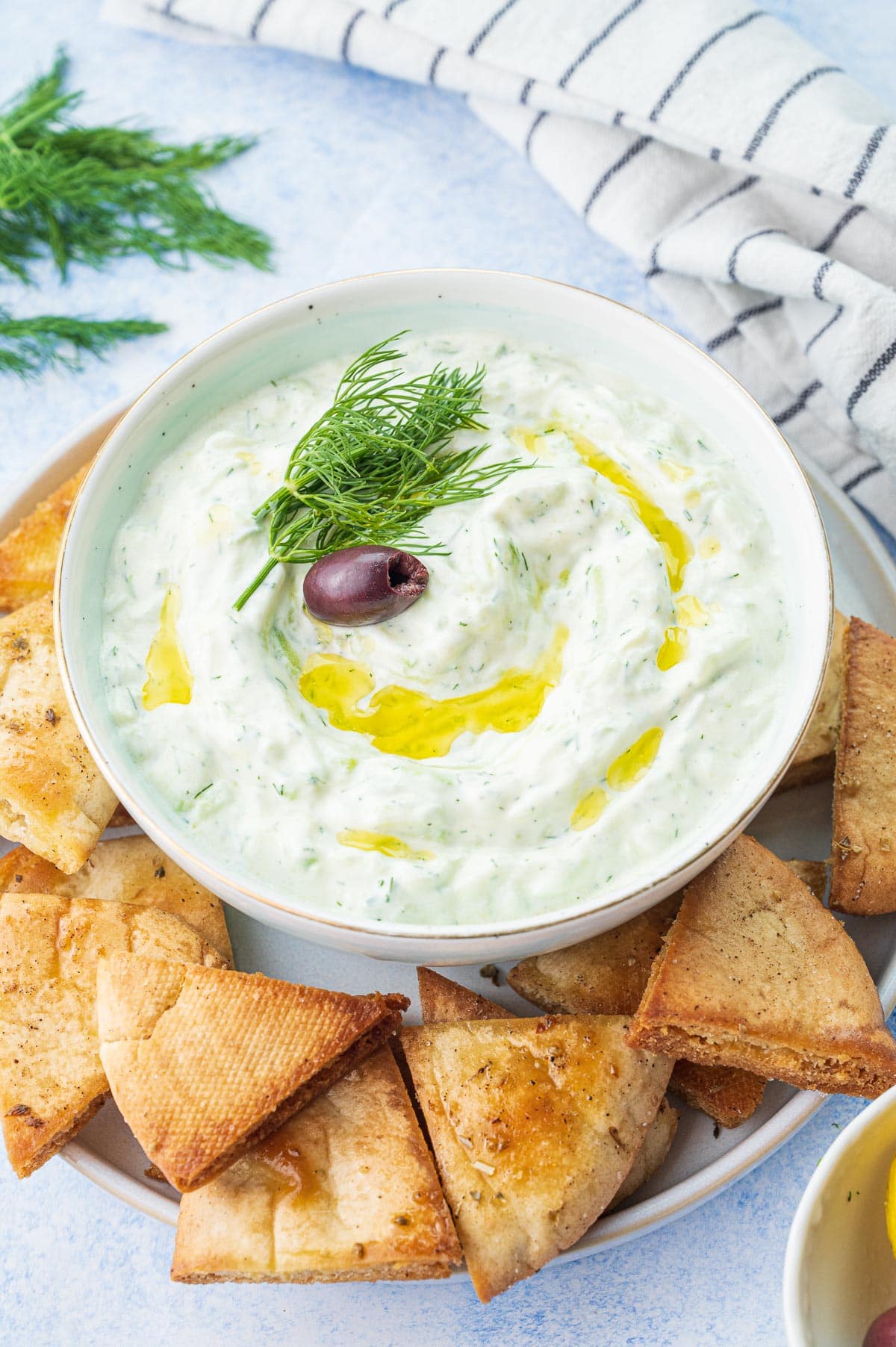 Tzatziki dip topped with dill, olive oil, and an olive in a white bowl.