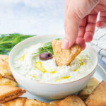 Tzatziki dip in a white bowl is being scooped with a pita chip.