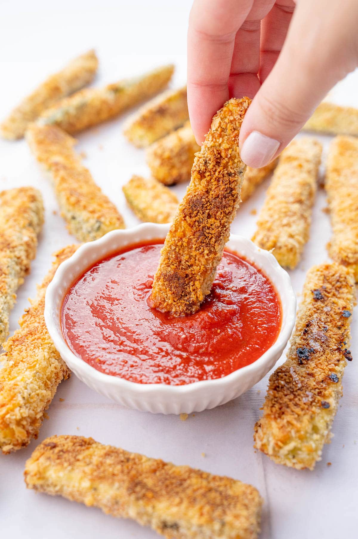 Zucchini fry is being dipped in marinara sauce. More zucchini fries scattered around.