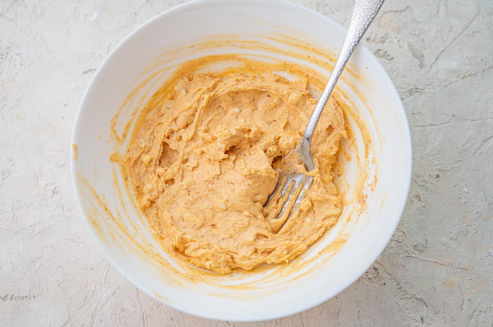 Cheese with spices mashed with a fork in a white bowl.