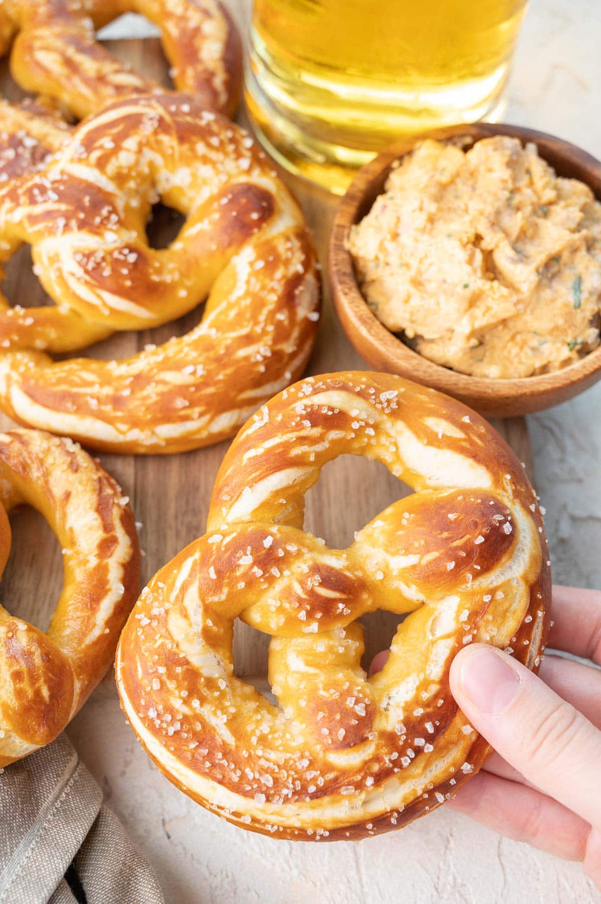 Soft pretzels on a wooden board. Beer and cheese dip on the side. One pretzel is held in a hand.