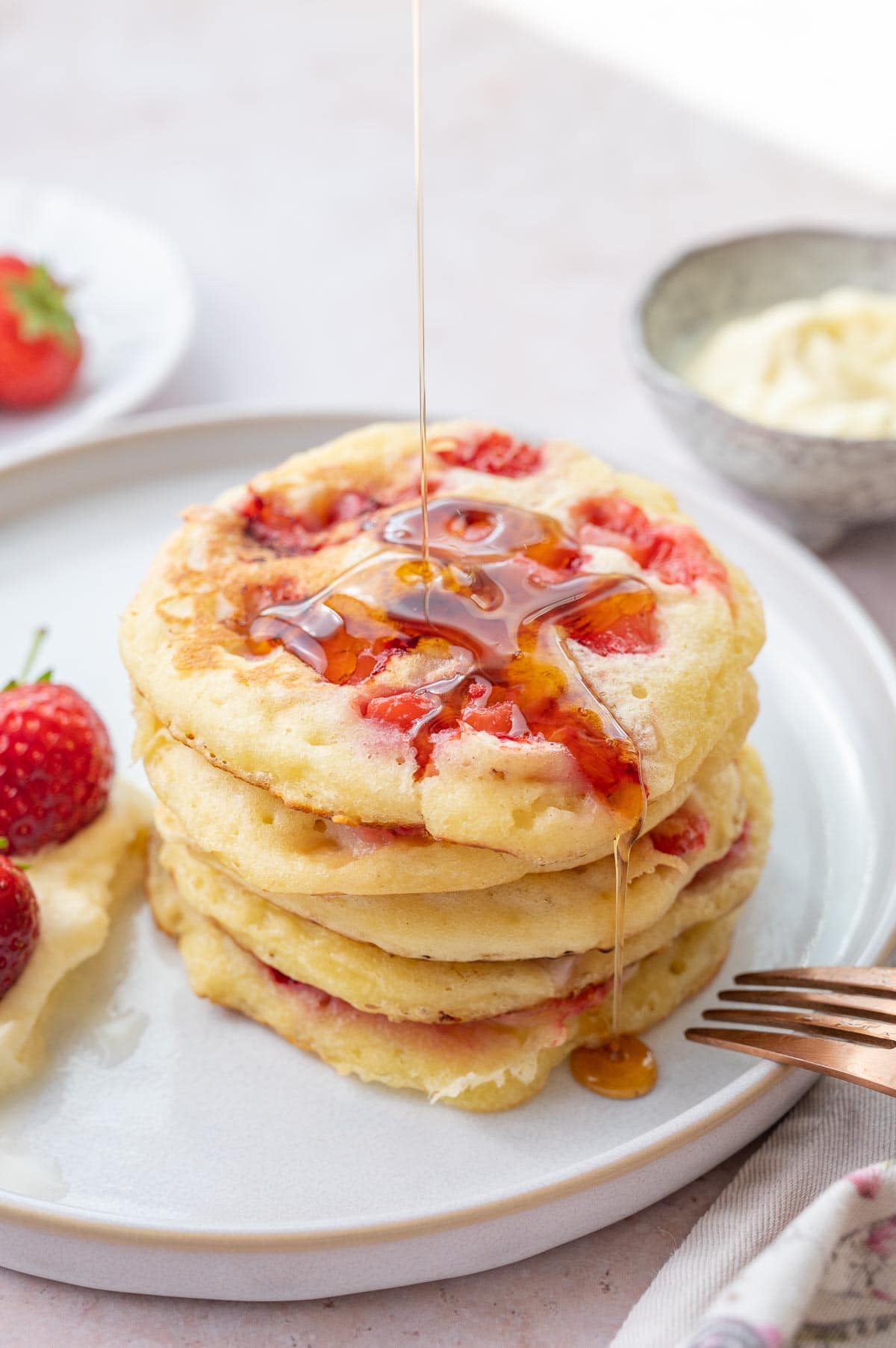 A stack of strawberry pancaked on a white plate poured with maple syrup.