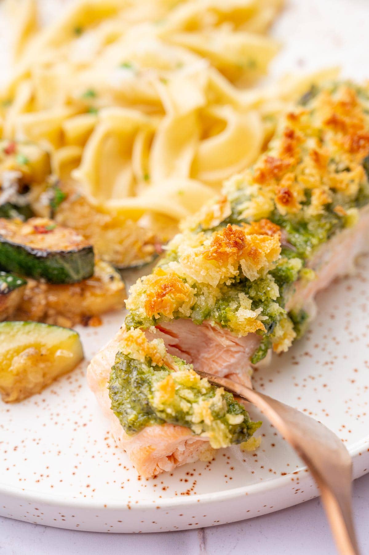 Baked pesto salmon on a white plate is being cut with a fork.