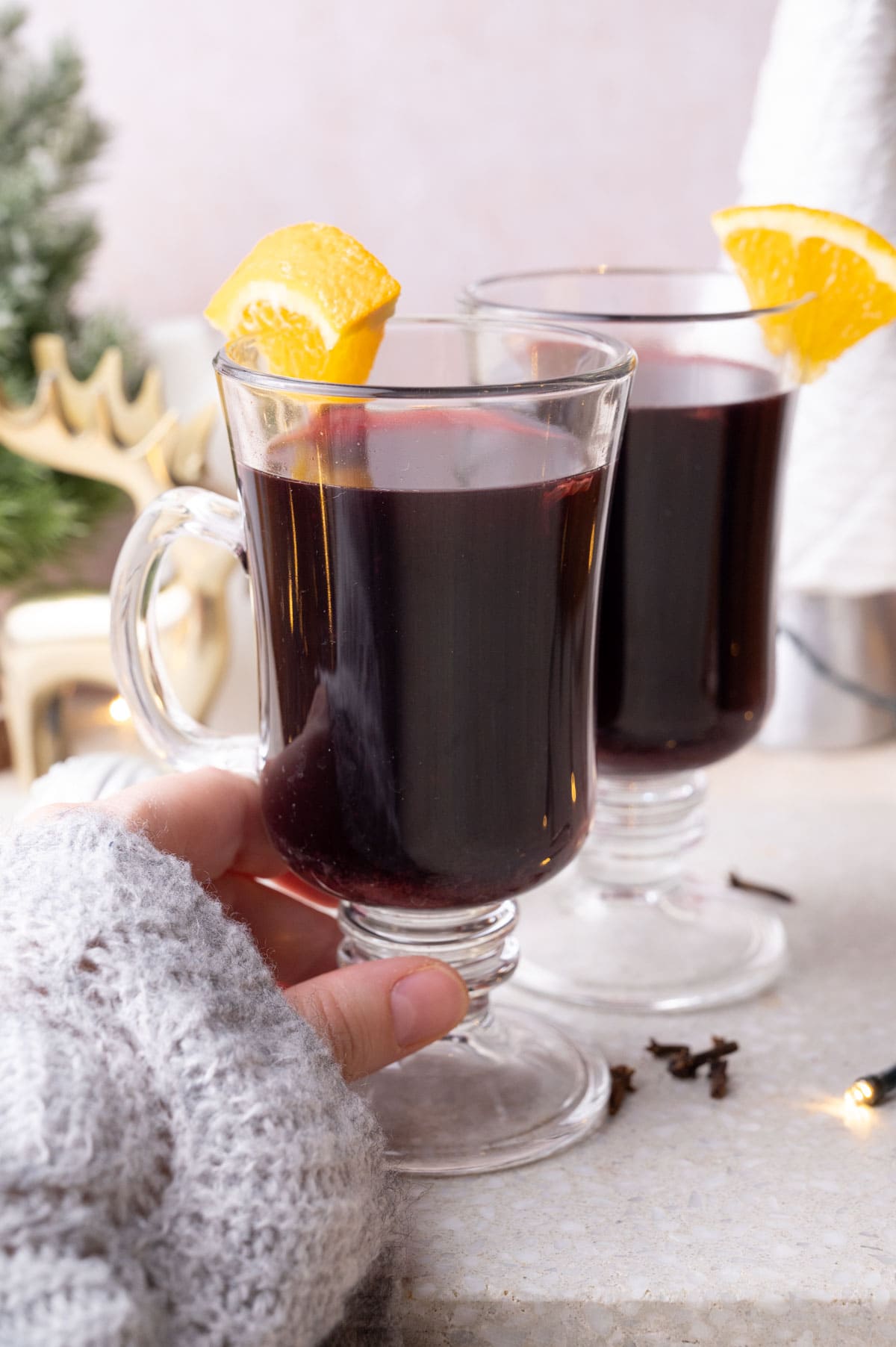 Two glasses with Glühwein on a grey stone board garnished with orange wedges. One glass is held in a hand.