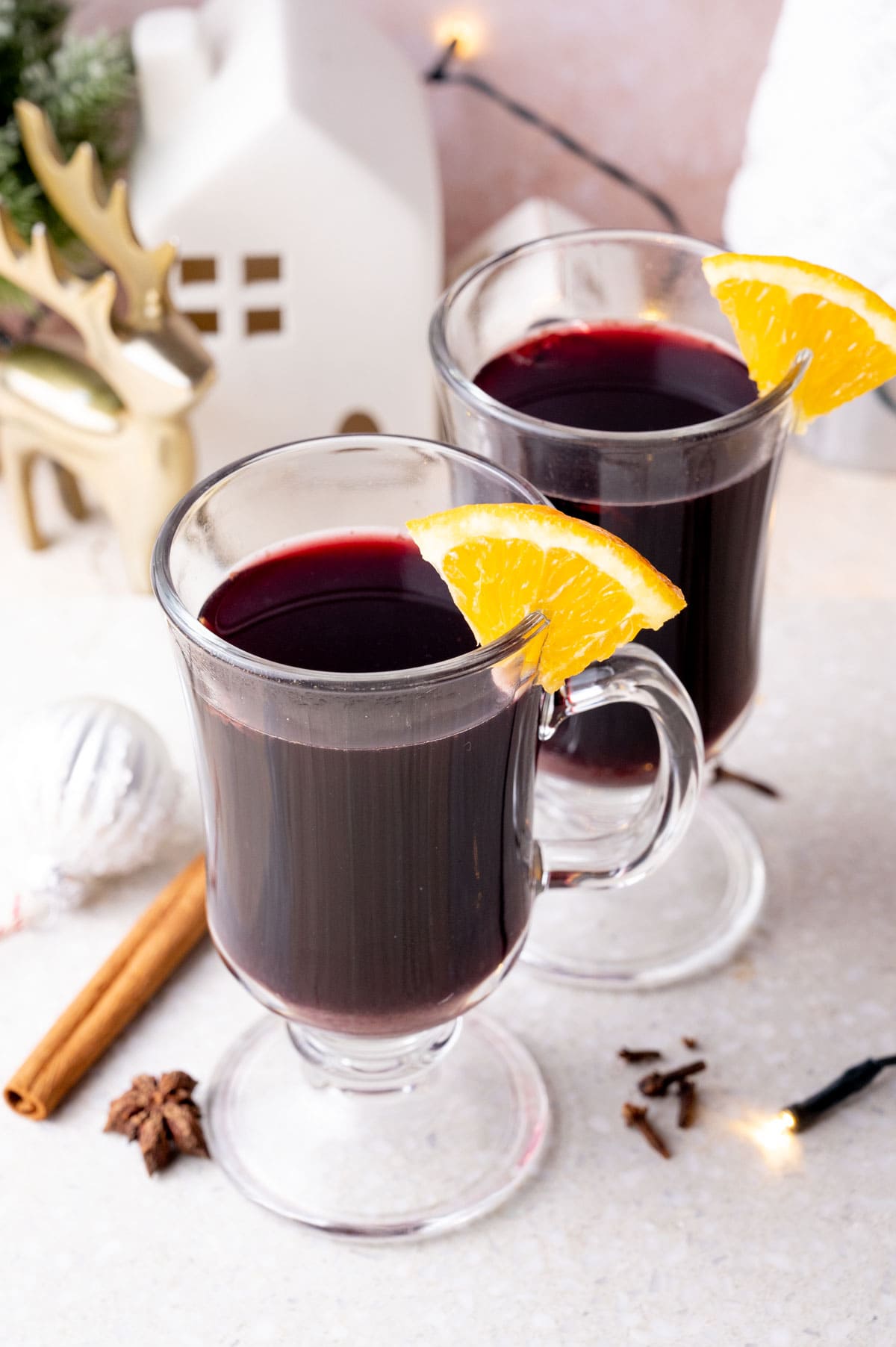 Two glasses with Glühwein on a grey stone board garnished with orange wedges. Christmas decorations in the background.