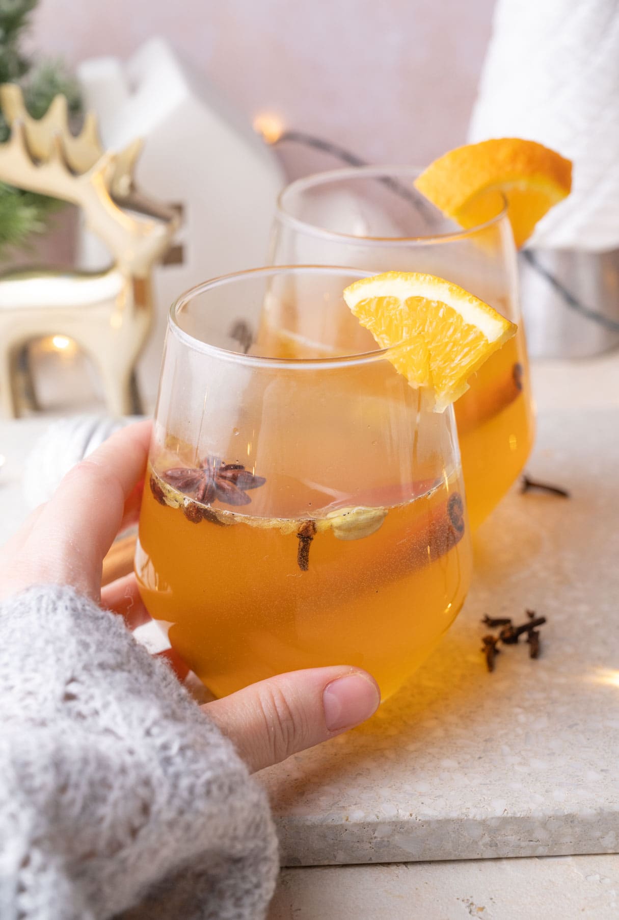 Two glassed with mulled white wine on a grey stone board. One glass is being held in a hand.