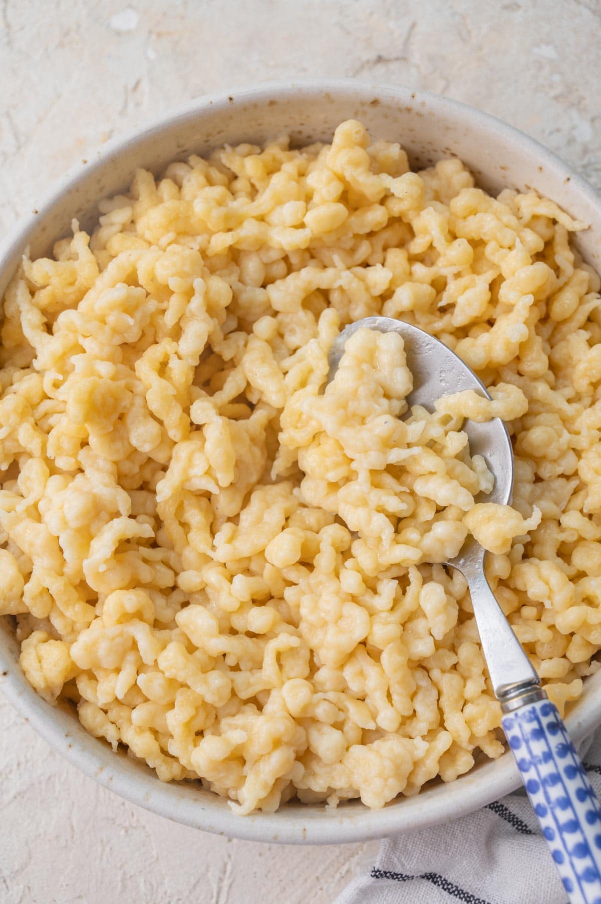 Cooked Spätzle in a beige bowl with blue spoon on the side.