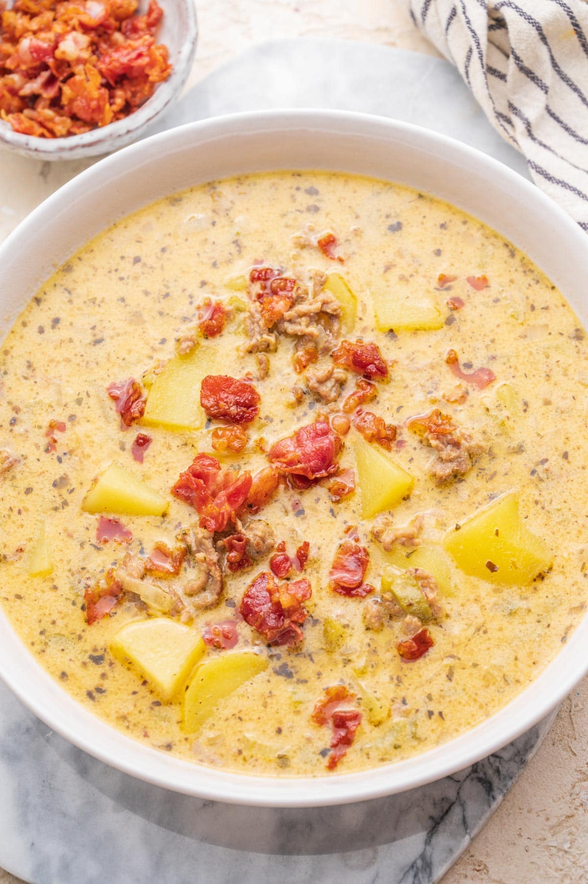 Cheeseburger chowder soup in a white bowl. A small bowl with bacon and a kitchen cloth on the side.