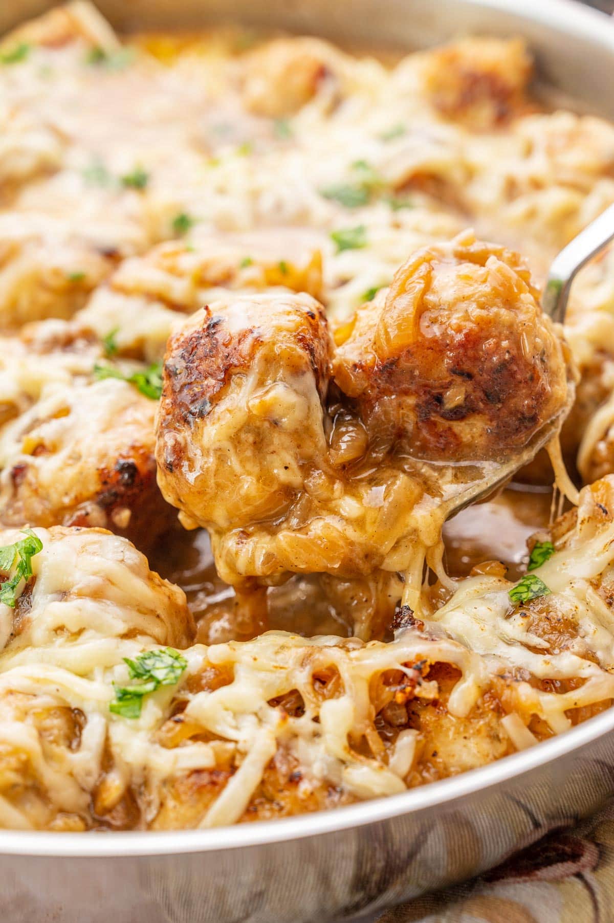 A close up photo of French onion meatballs in a pan. Two meatballs with melted cheese are on a spoon.
