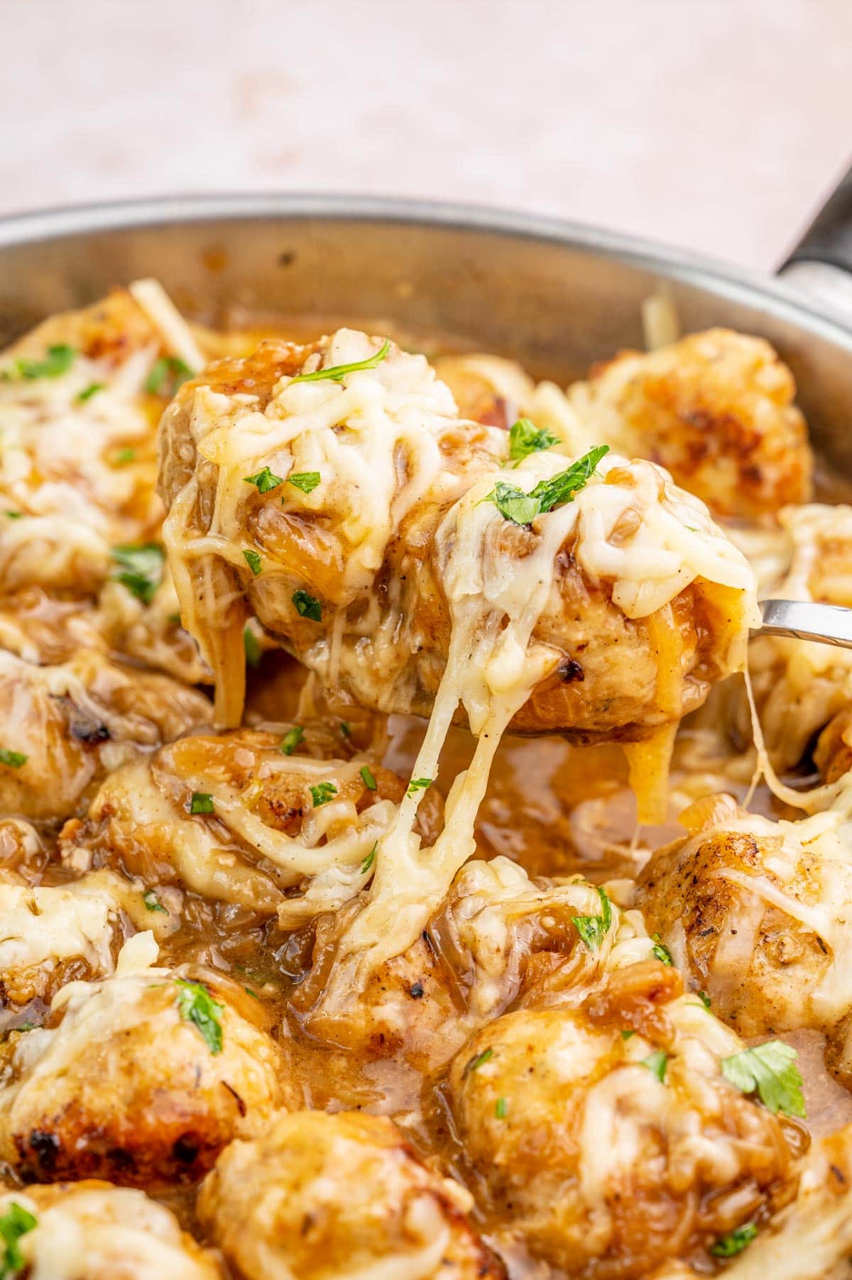 A close up photo of French onion meatballs in a pan. Two meatballs with melted cheese are on a spoon.