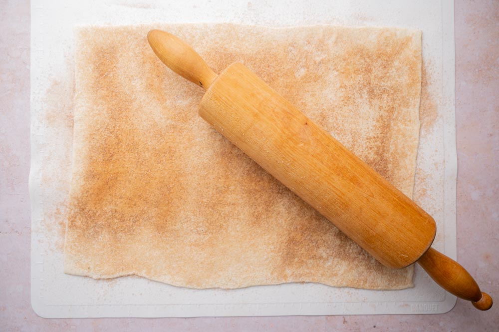 Puff pastry sheet topped with cinnamon sugar is being rolled out with a mallet.