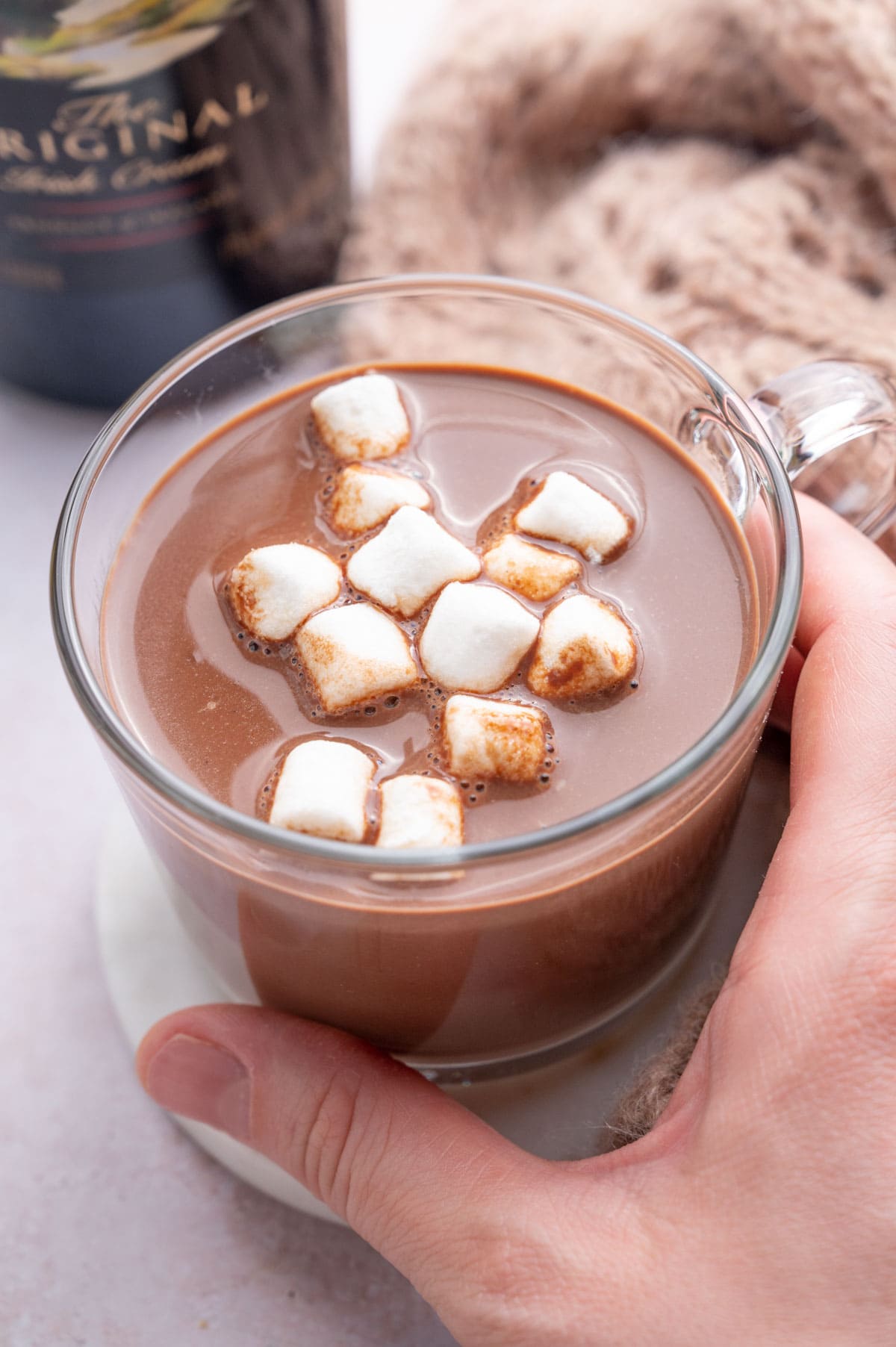 Baileys hot chocolate in a glass topped with marshmallows. A glass is being held in a hand.
