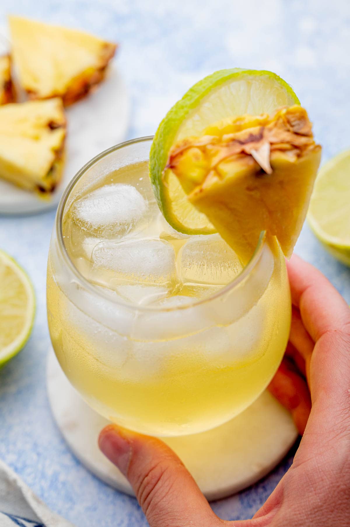 A glass with pineapple vodka drink held in a hand. A blue background with pineapple wedges and limes.