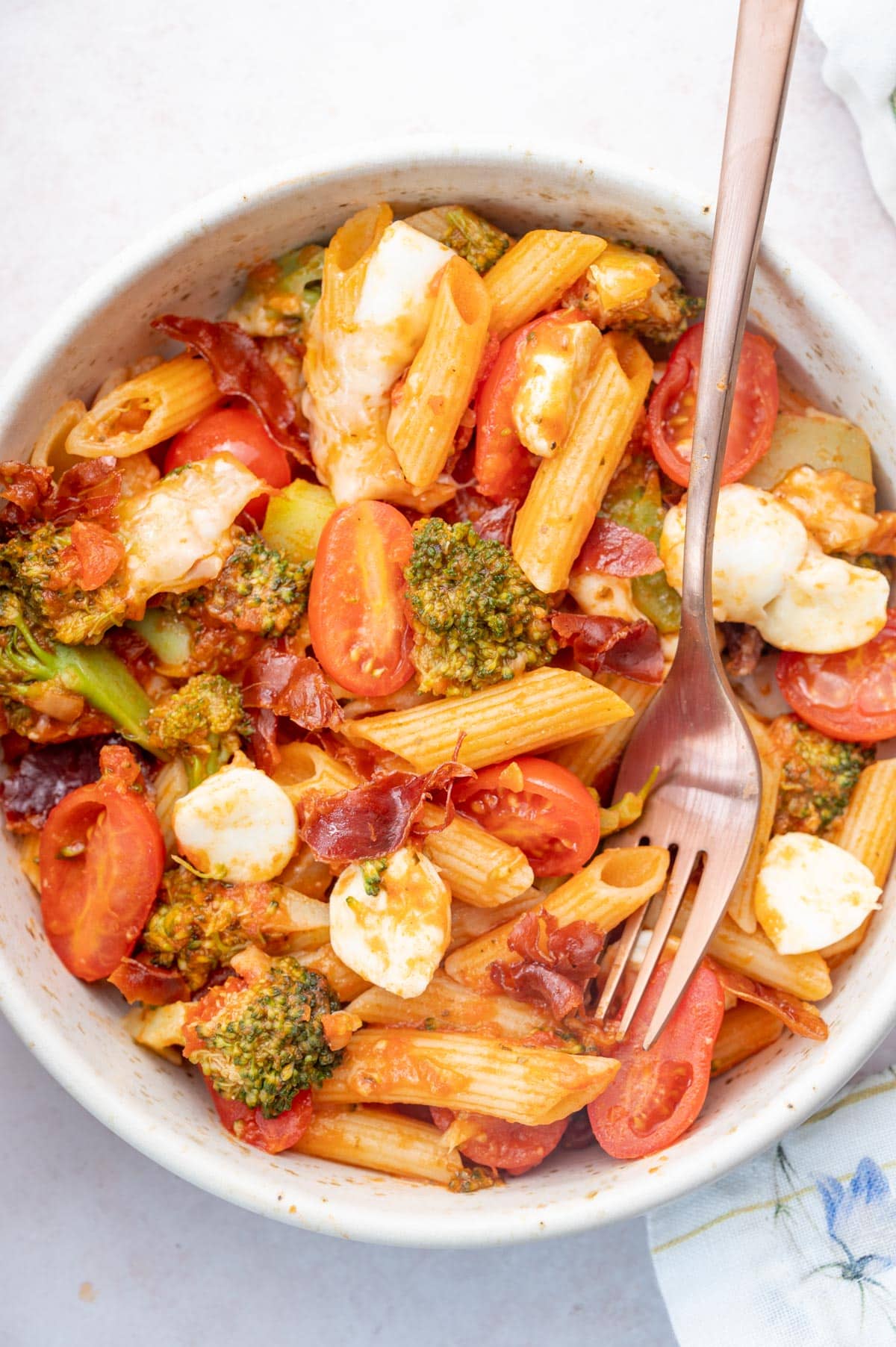 Cheesy Tomato Broccoli Pasta with Crispy Prosciutto in a white bowl with a rose gold fork on the side.