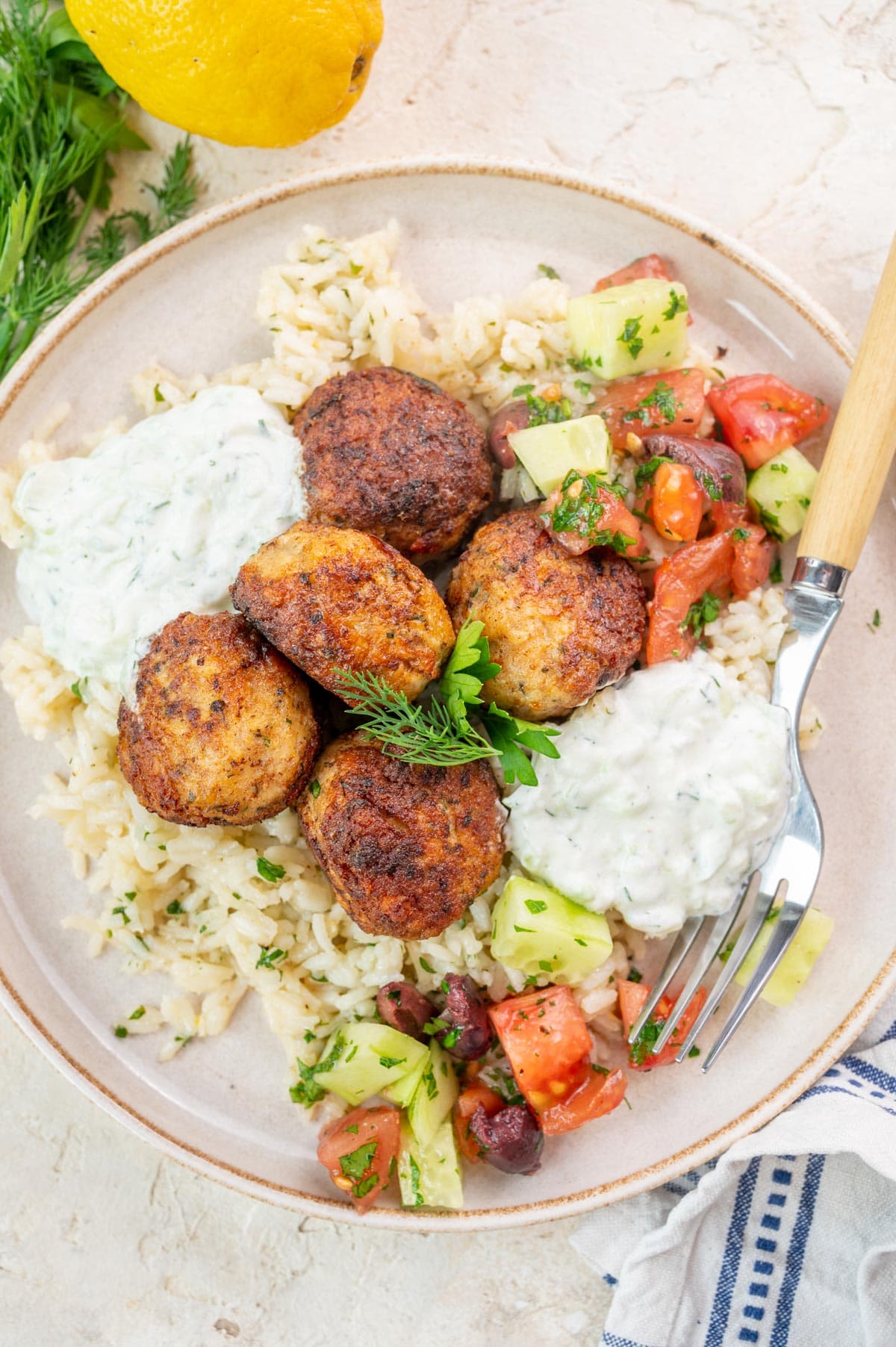 Greek Chicken Meatballs with rice, tzatziki and tomato cucumber salad on a white plate.