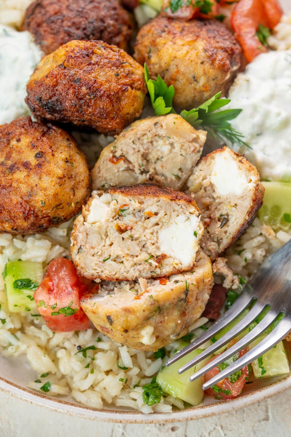 A close up photo of Greek Chicken Meatballs with rice and tomato cucumber salad on a plate. Meatballs are cut in half to show the inside.