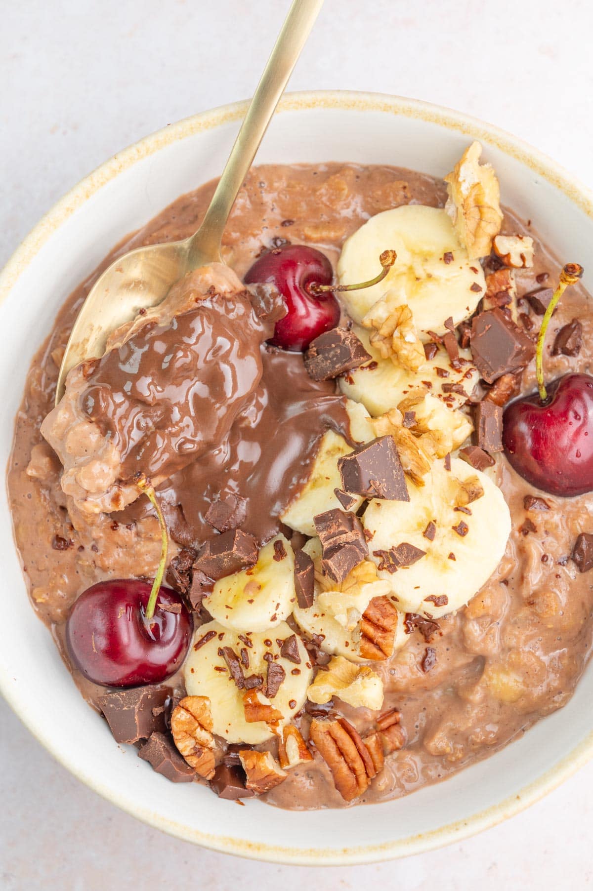 Banana chocolate oatmeal in a white bowl topped with banana slices, chocolate chunks, and cherries.