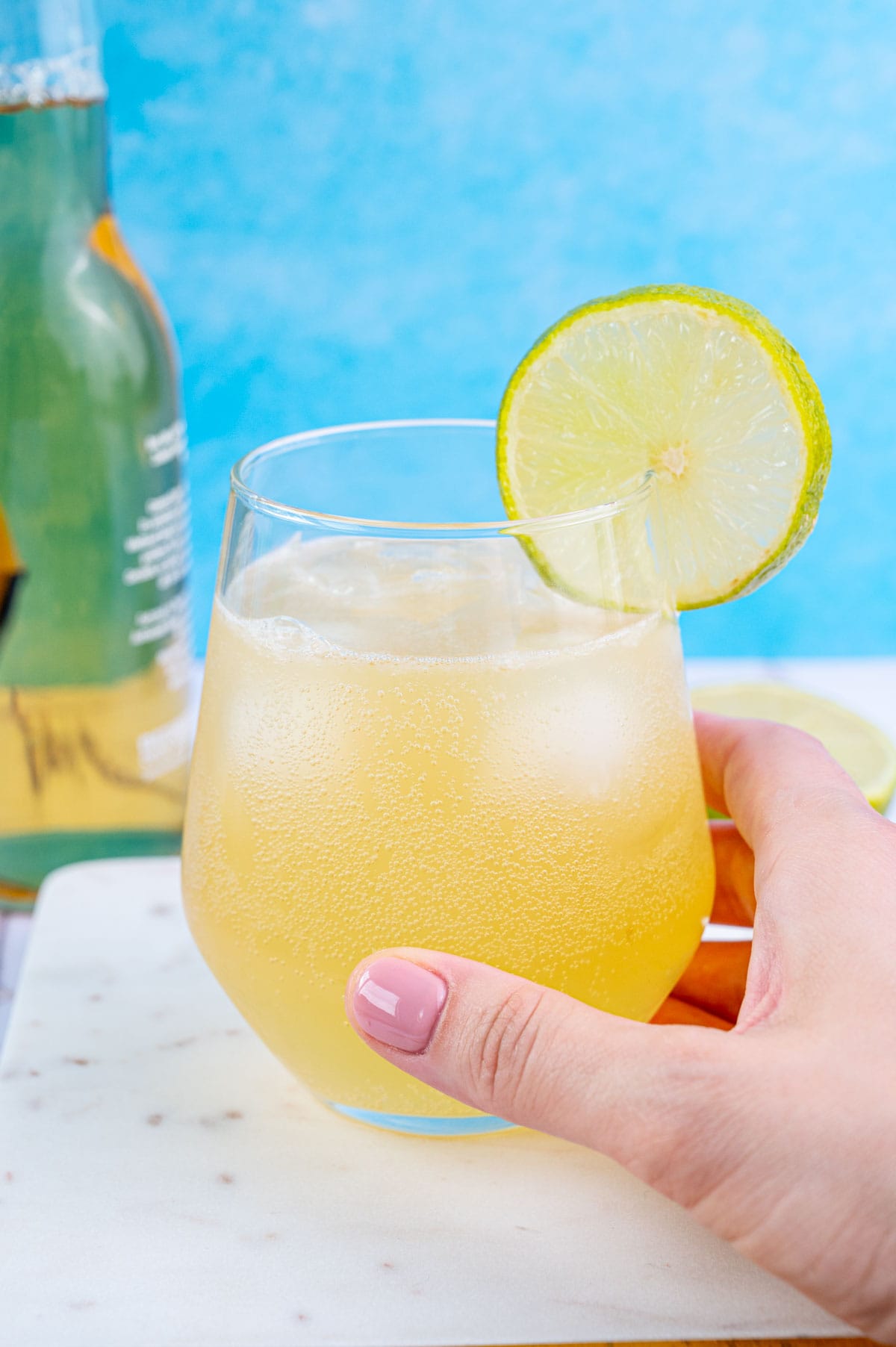 Beer Margarita in a short glass held in a hand, garnished with lime slice on a blue background.