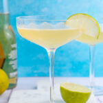 Beer Margarita in a coupe glass garnishes with a lime slice on a blue background.