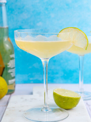 Beer Margarita in a coupe glass garnishes with a lime slice on a blue background.