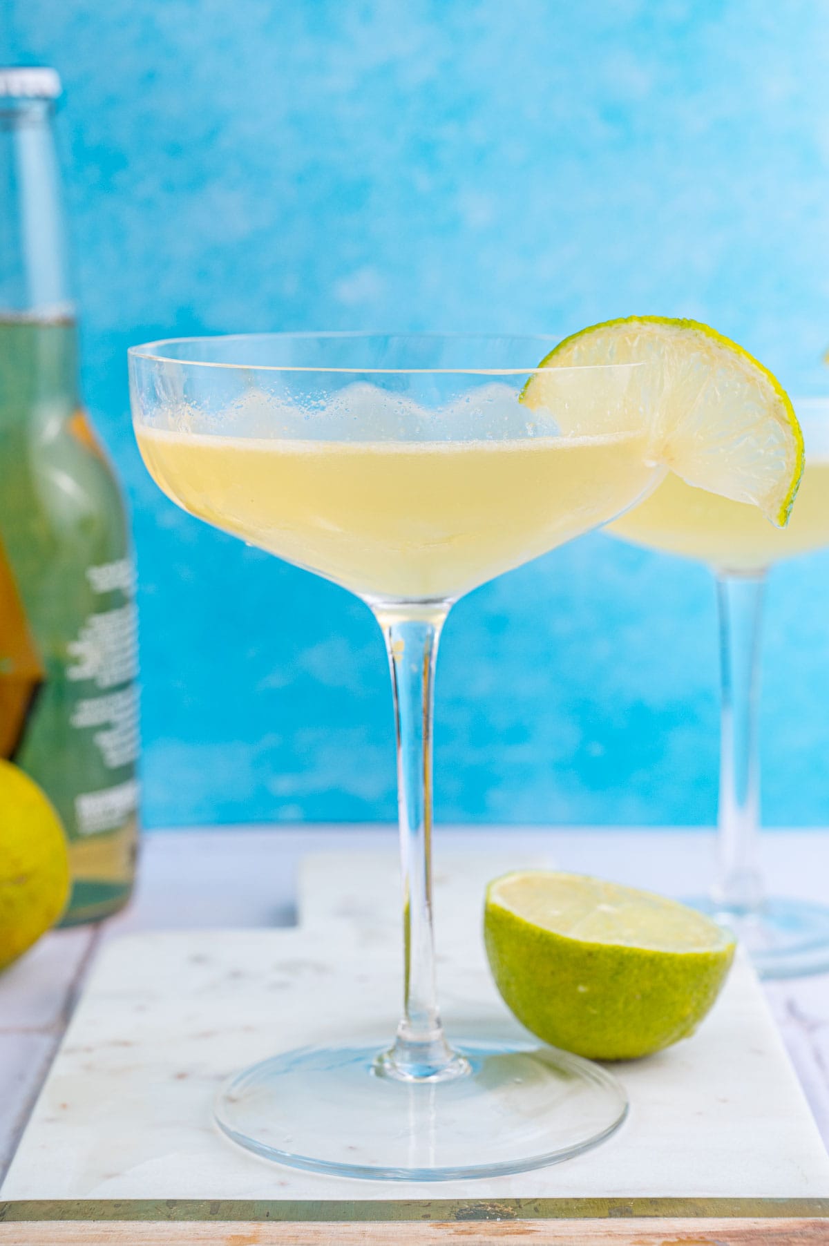 Beer Margarita in a coupe glass garnishes with a lime slice on a blue background.