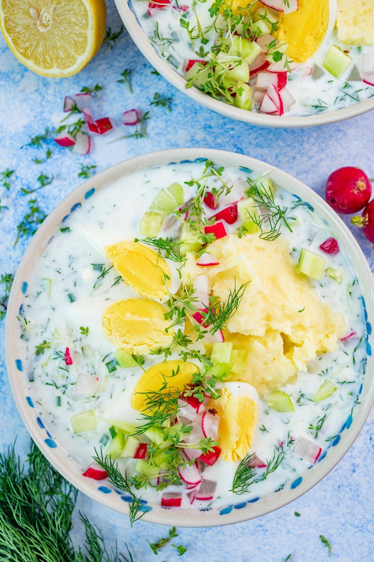 Two bowls with chlodnik ogorkowy soup topped with eggs, potatoes, and radishes on a blue background.