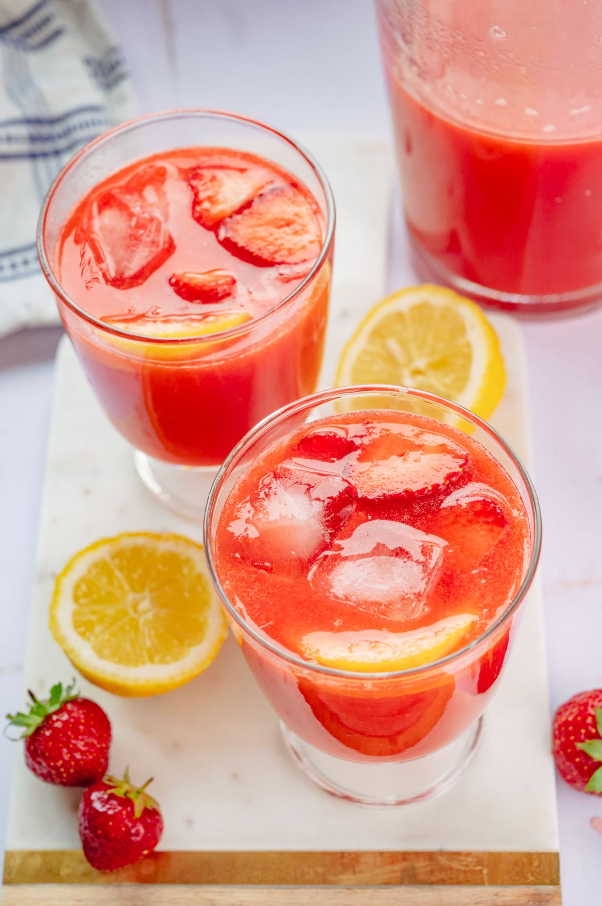 Two glasses with strawberry lemonade and ice cubes. Strawberries and lemons in the background.