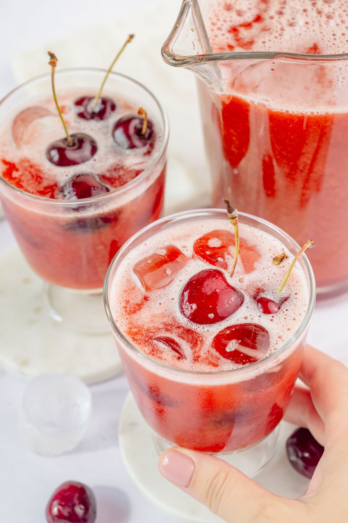 Two glasses and a pitcher with cherry lemonade. One glass is held in a hand.