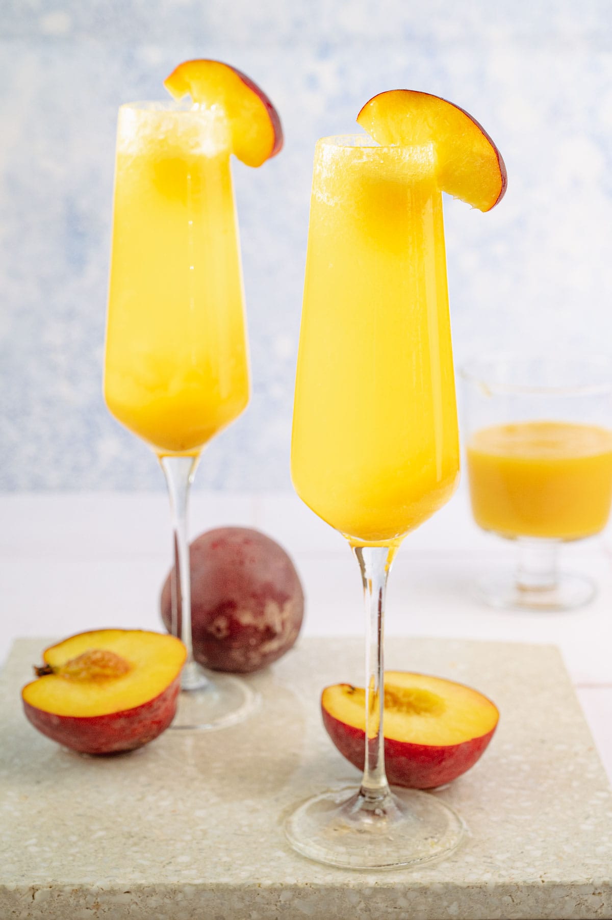 Two champagne glasses with peach bellini decorated with peach slices on a stone board.