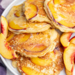 Peach pancakes on a white plate with fresh peach slices on the side.