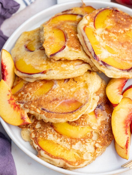 Peach pancakes on a white plate with fresh peach slices on the side.