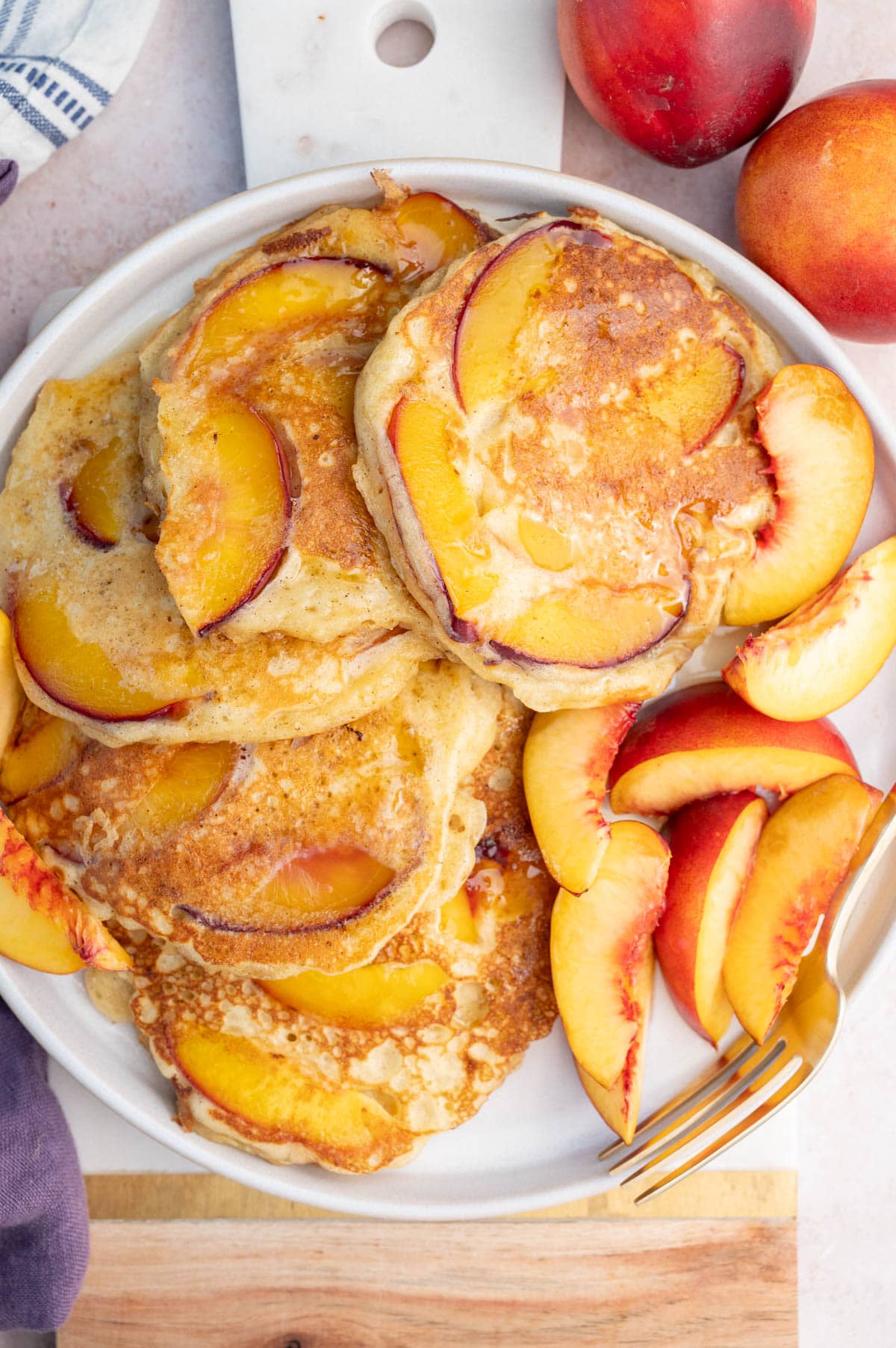 Peach pancakes on a white plate with fresh peach wedges. Whole peaches in the background.