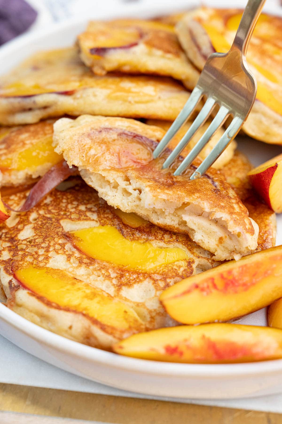 A close-up photo of peach pancakes on a white plate. A piece of a pancakes is being picked on a fork.