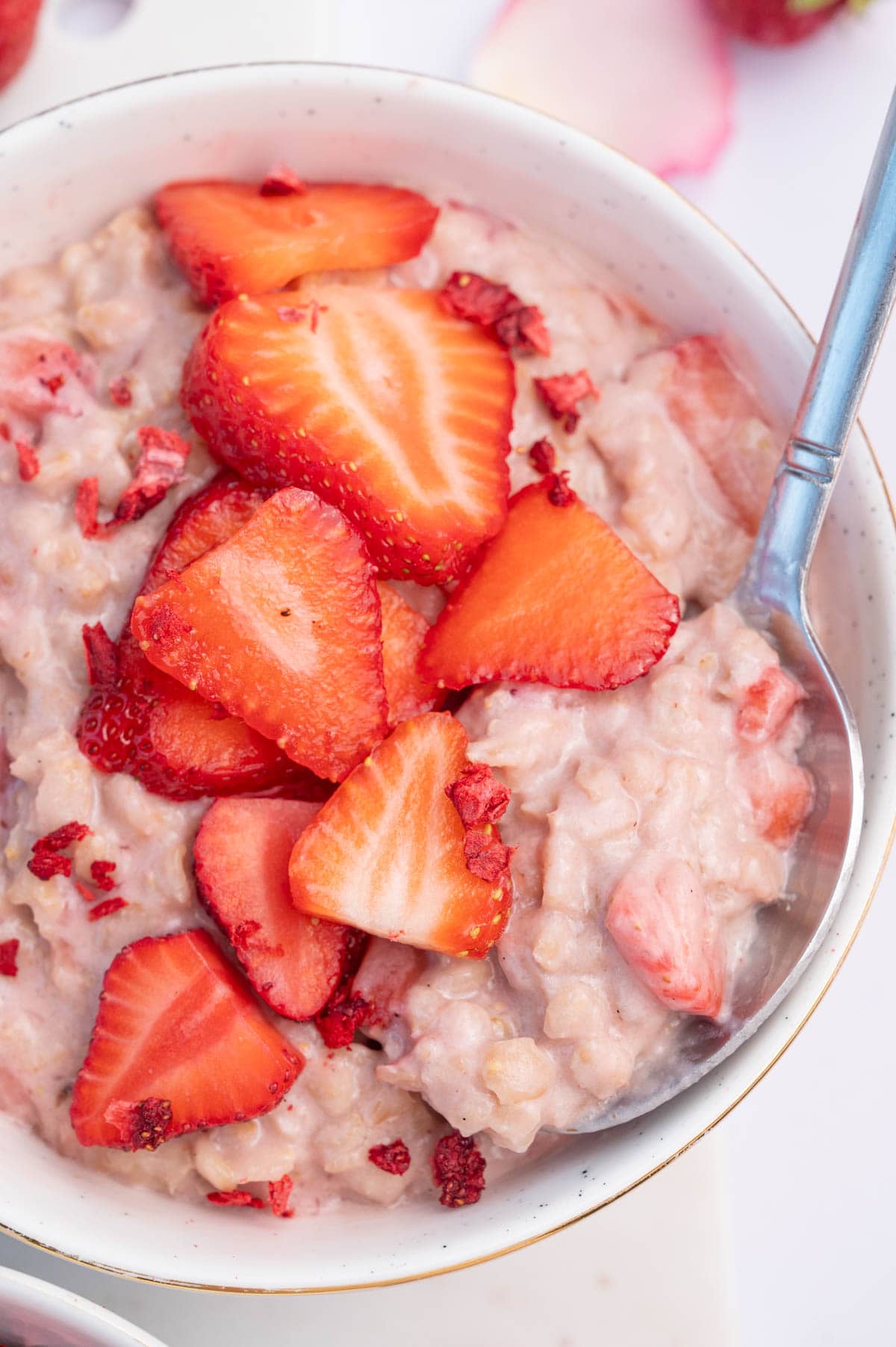 A close-up photo of strawberries and cream oatmeal in a white bowl topped with slices strawberries with a spoon on the side.