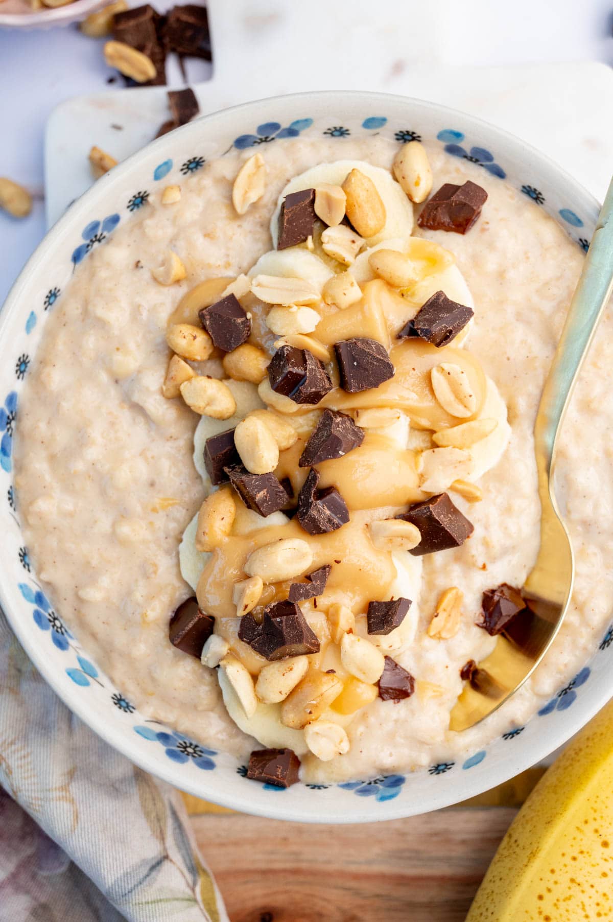 Banana peanut butter oatmeal in a white-blue bowl topped with banana slices, chocolate, and peanuts.