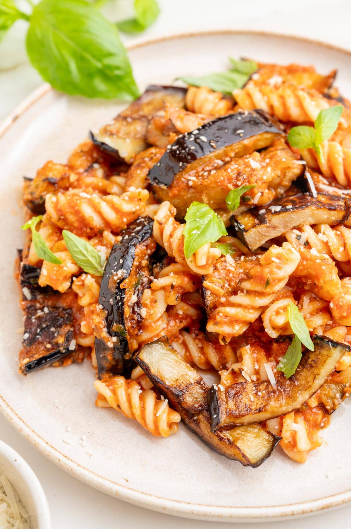 Pasta alla Norma on a white plate sprinkled with parmesan and basil leaves.