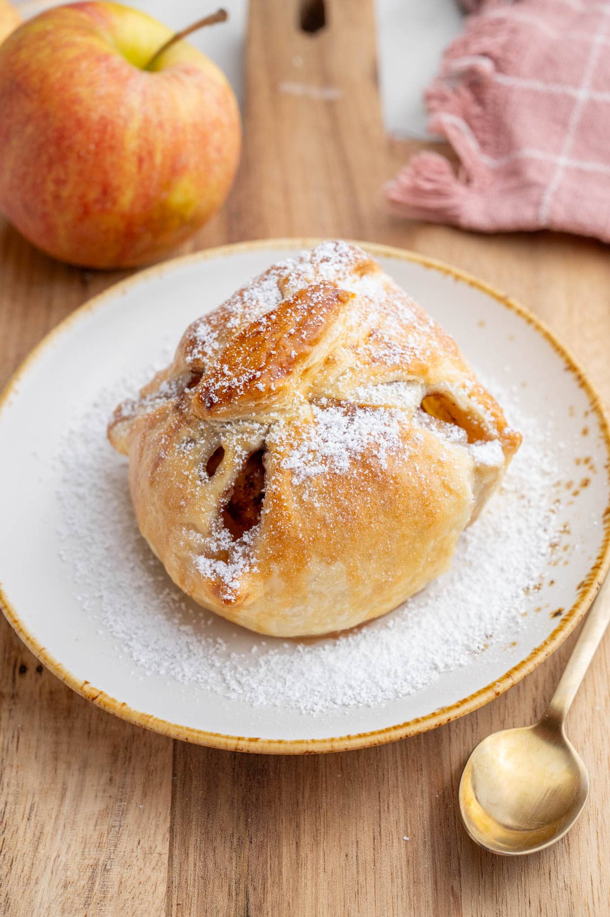 An apple in puff pastry sprinkled with powdered sugar on a white plate on a wooden board.