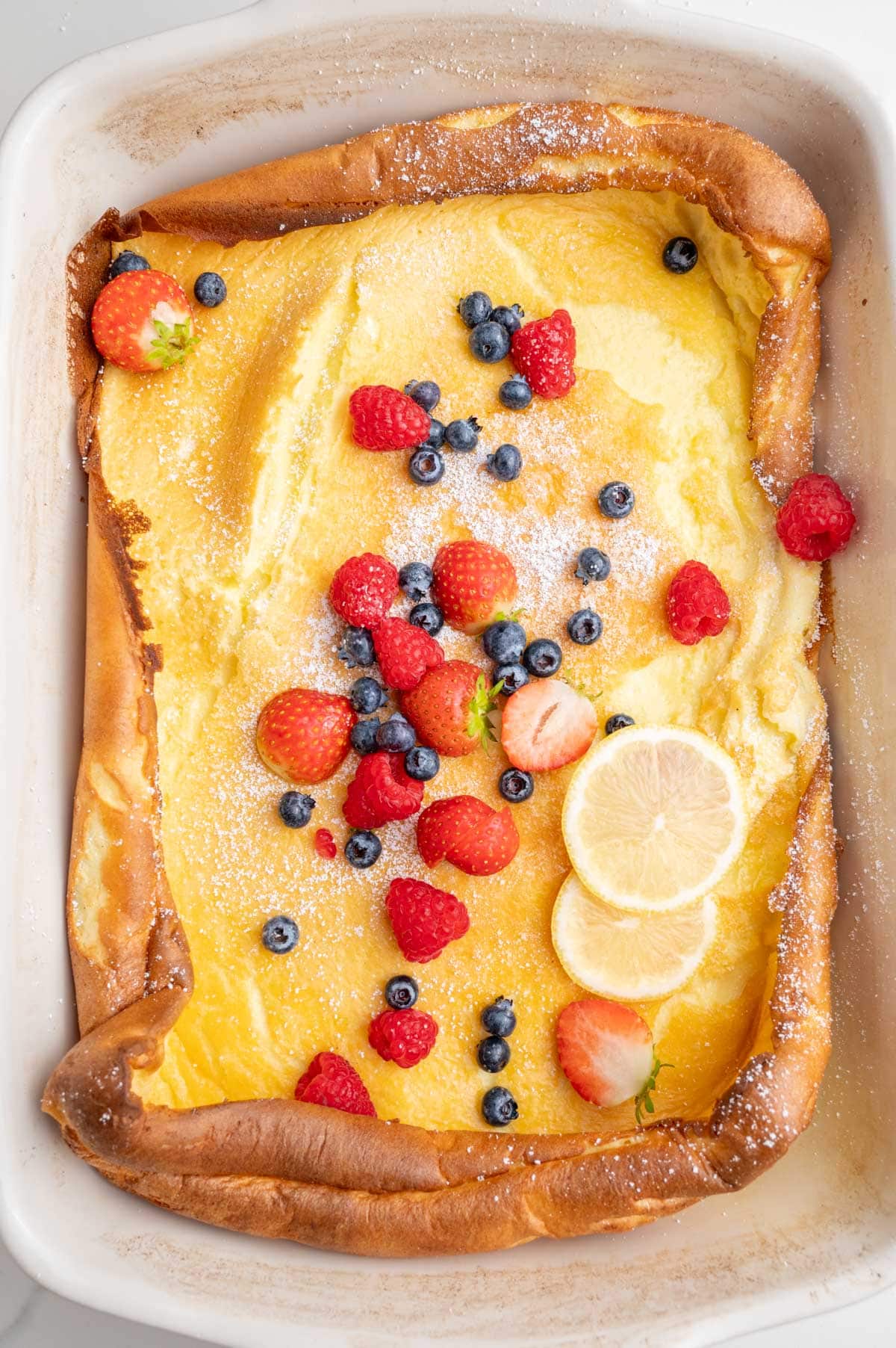 Dutch baby pancake in a white casserole dish topped with berries and lemon slices.