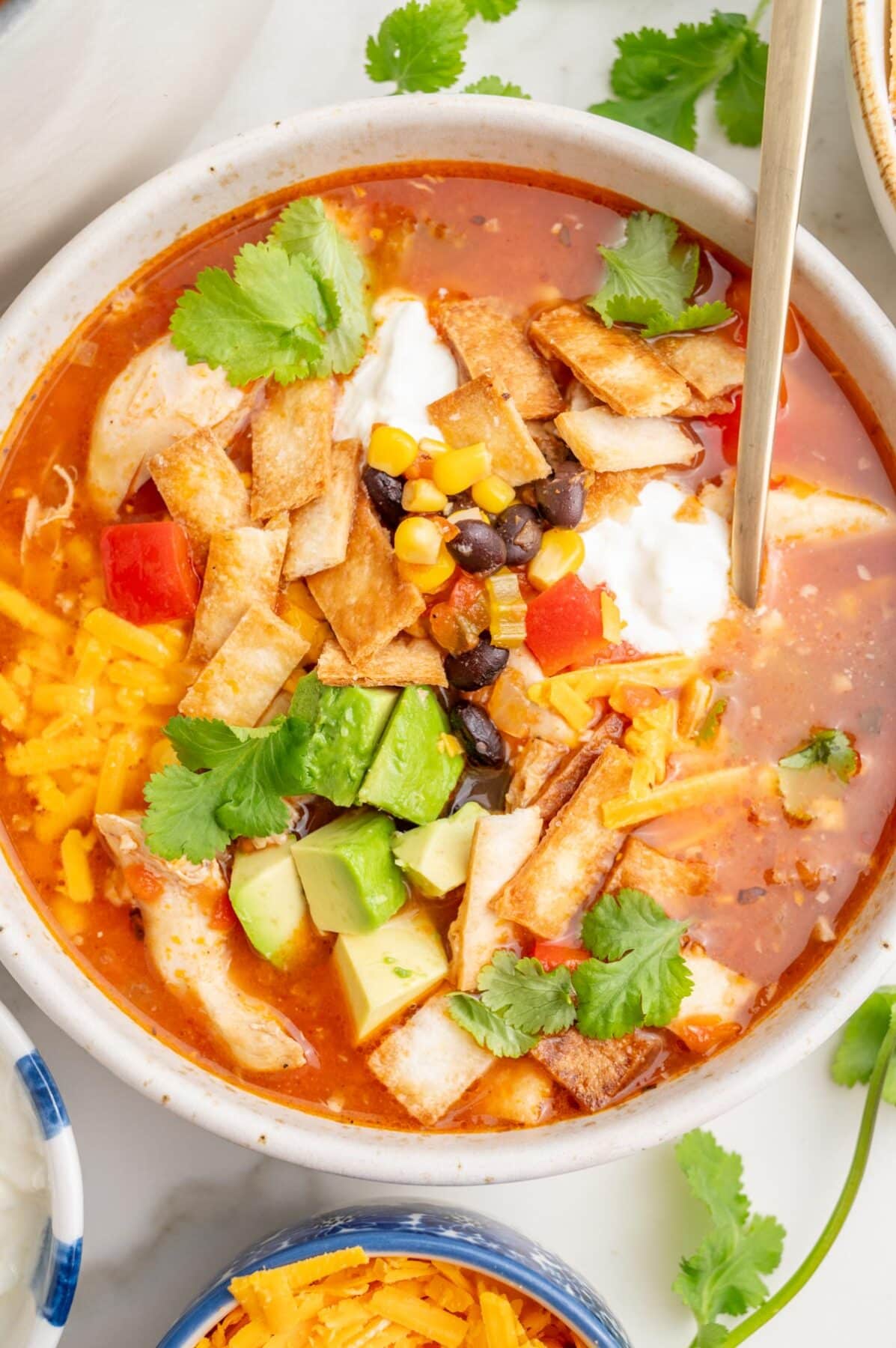 An overhead photo of chicken tortilla soup in a white bowl topped with avocado, tortilla strips, cheddar, and cilantro.