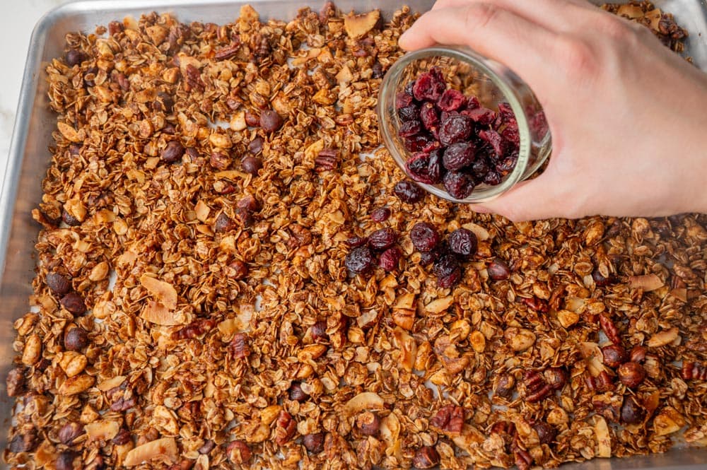 Dried cranberries are being added to baked gingerbread granola on a baking sheet.