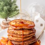 Orange gingerbread syrup is being poured over a stack of gingerbread pancakes on a white plate.